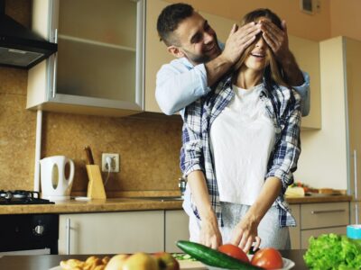 Man playfully covers woman's eyes in kitchen.