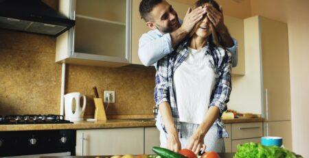 Man playfully covers woman's eyes in kitchen.